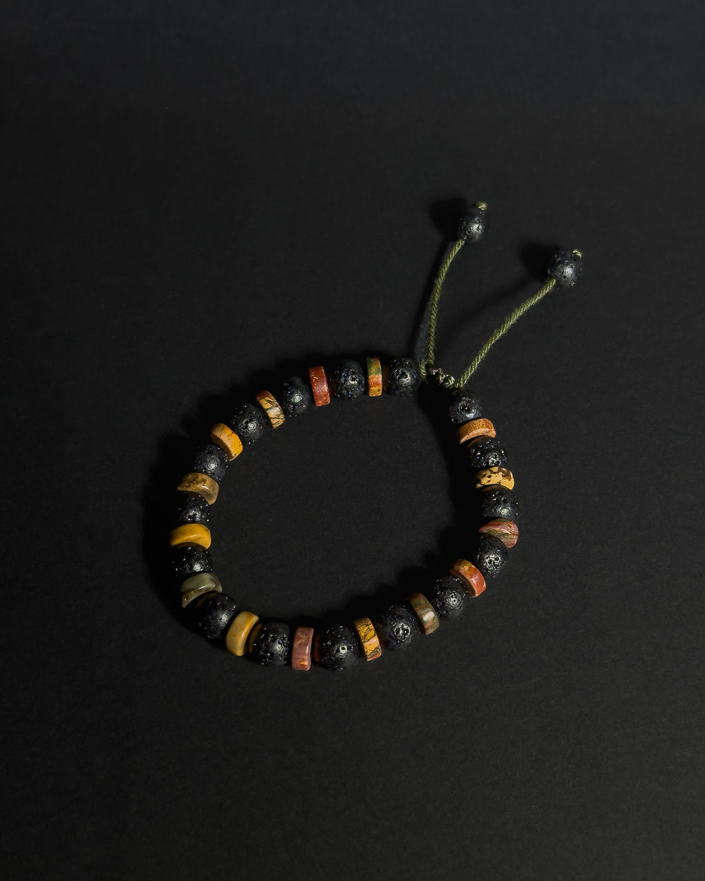 A beaded adjustable bracelet featuring black lava stones, and picasso jasper, with an adjustable green cord closure, displayed on a dark background.