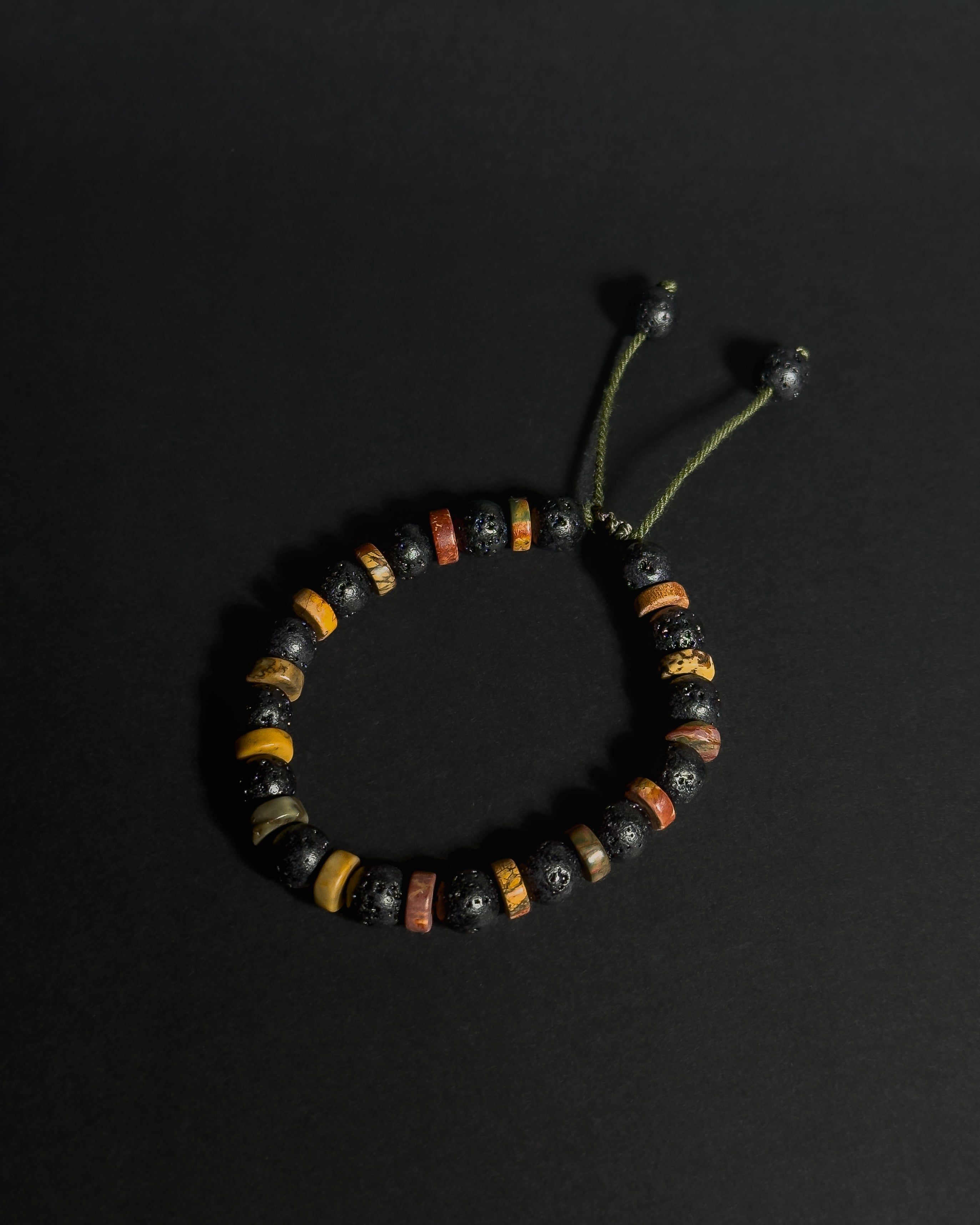A beaded adjustable bracelet featuring black lava stones, and picasso jasper, with an adjustable green cord closure, displayed on a dark background.