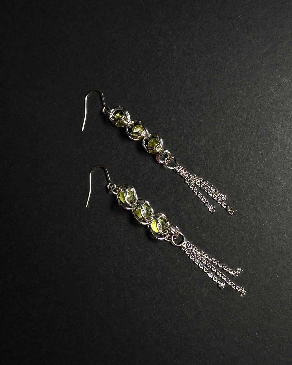 A pair of silver dangle earrings featuring three faceted light green gemstones, peridot in a wrapped capture bead setting, with three delicate silver chain tassels hanging from each earring, displayed on a dark textured background.