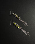 A pair of silver dangle earrings featuring three faceted light green gemstones, peridot in a wrapped capture bead setting, with three delicate silver chain tassels hanging from each earring, displayed on a dark textured background.