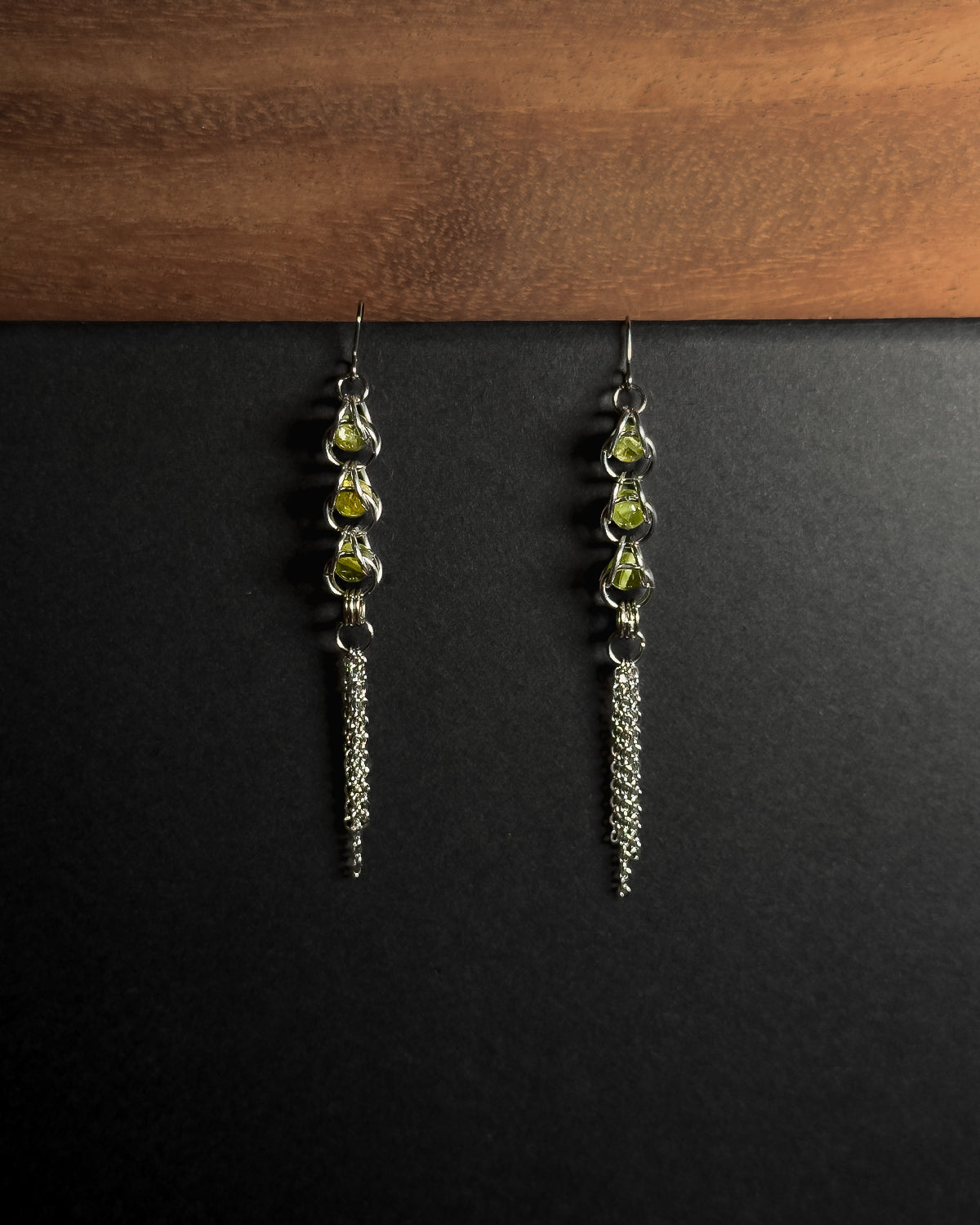 A pair of silver-toned dangle earrings with three small green peridot gemstones and a long dangling chain, displayed against a black background with a wooden surface at the top.