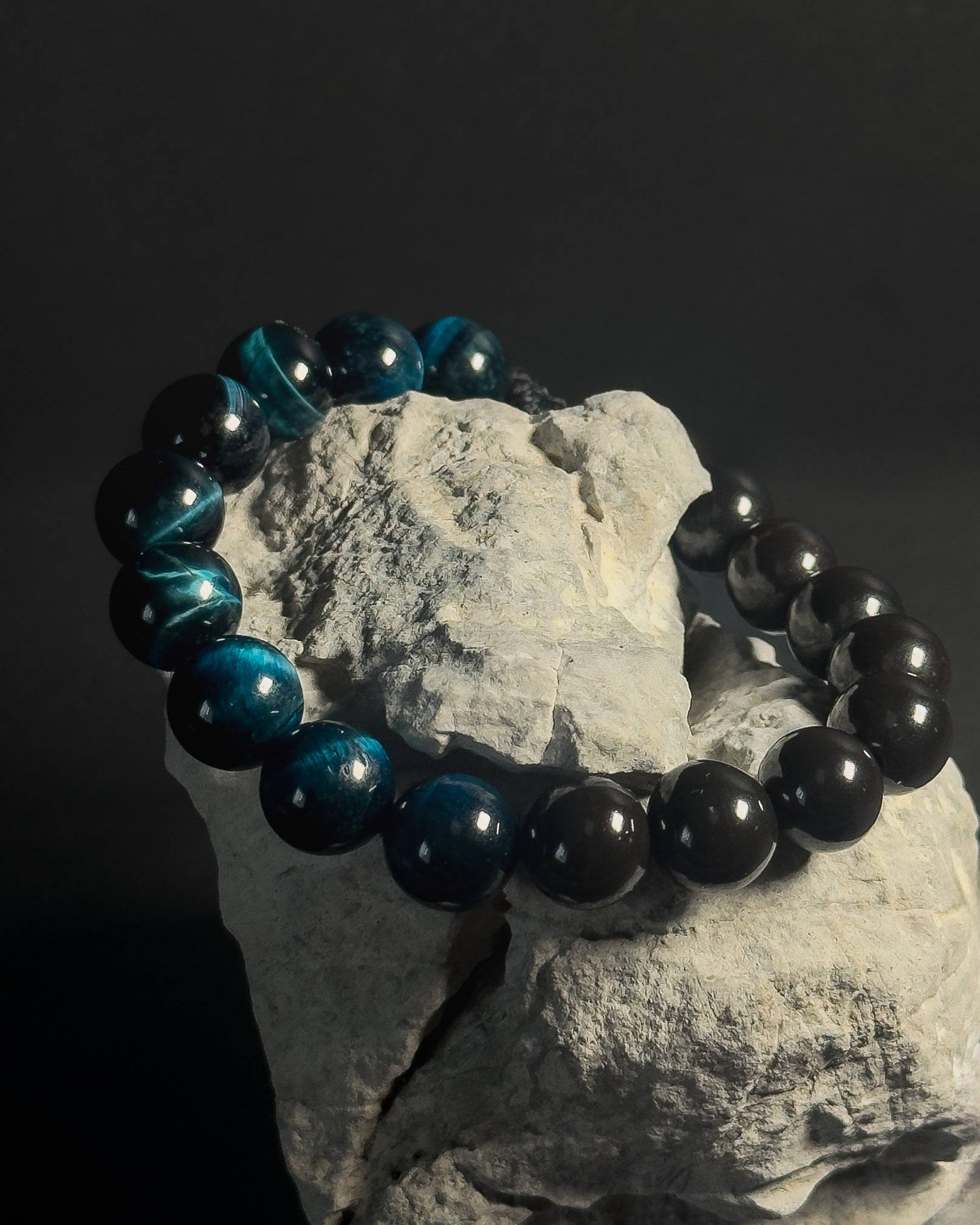Black and blue beaded bracelet on a rock with a dark background. Black beaded bracelet with blue tiger eye.