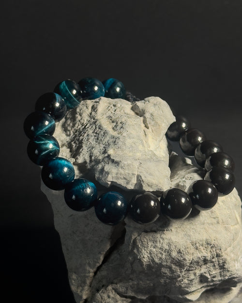 Black and blue beaded bracelet on a rock with a dark background. Black beaded bracelet with blue tiger eye.