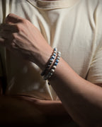 A close-up of a person's wrist wearing two beaded bracelets, made with natural K2 stone, against a cream-colored t-shirt.