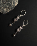 A pair of silver dangle earrings with rose quartz crystal capture bead and dark gray pearl drops, displayed on a black background with a textured gray stone prop.
