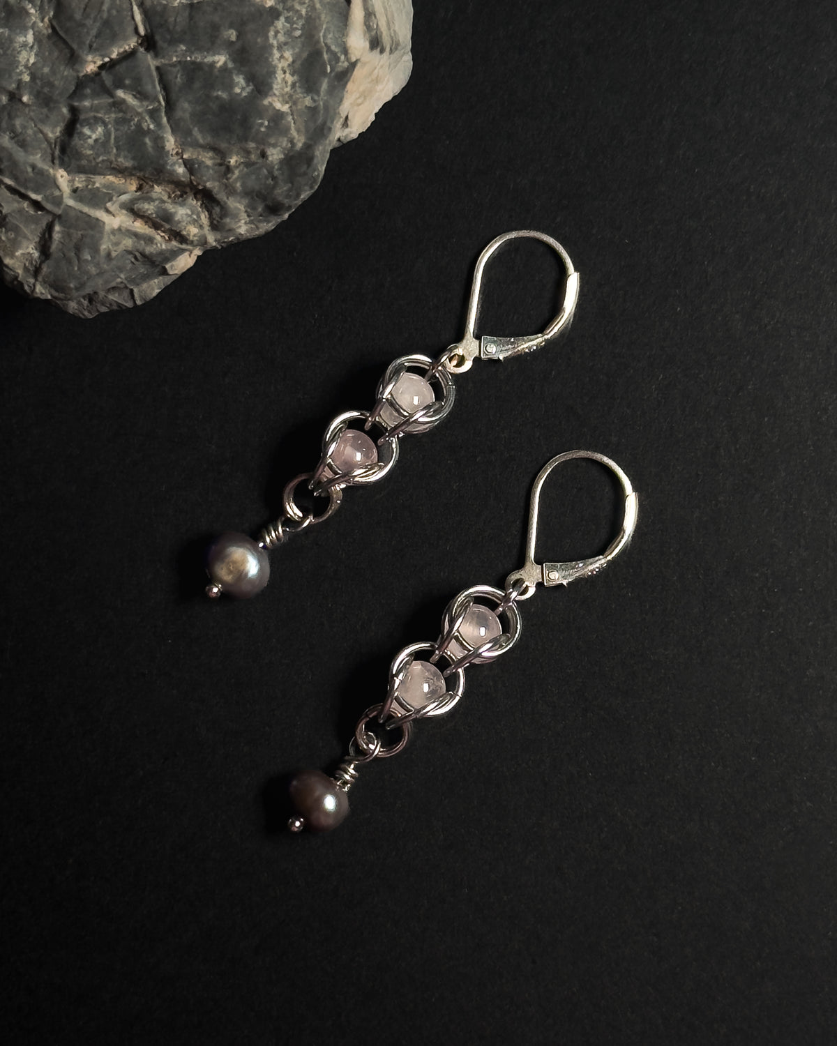 A pair of silver dangle earrings with rose quartz crystal capture bead and dark gray pearl drops, displayed on a black background with a textured gray stone prop.