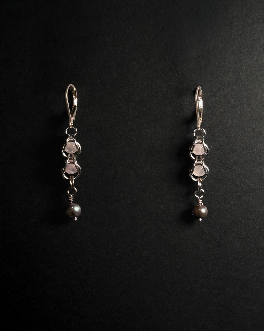 A pair of silver dangle earrings with rose quartz crystal capture bead and dark gray pearl drops, displayed on a black background .