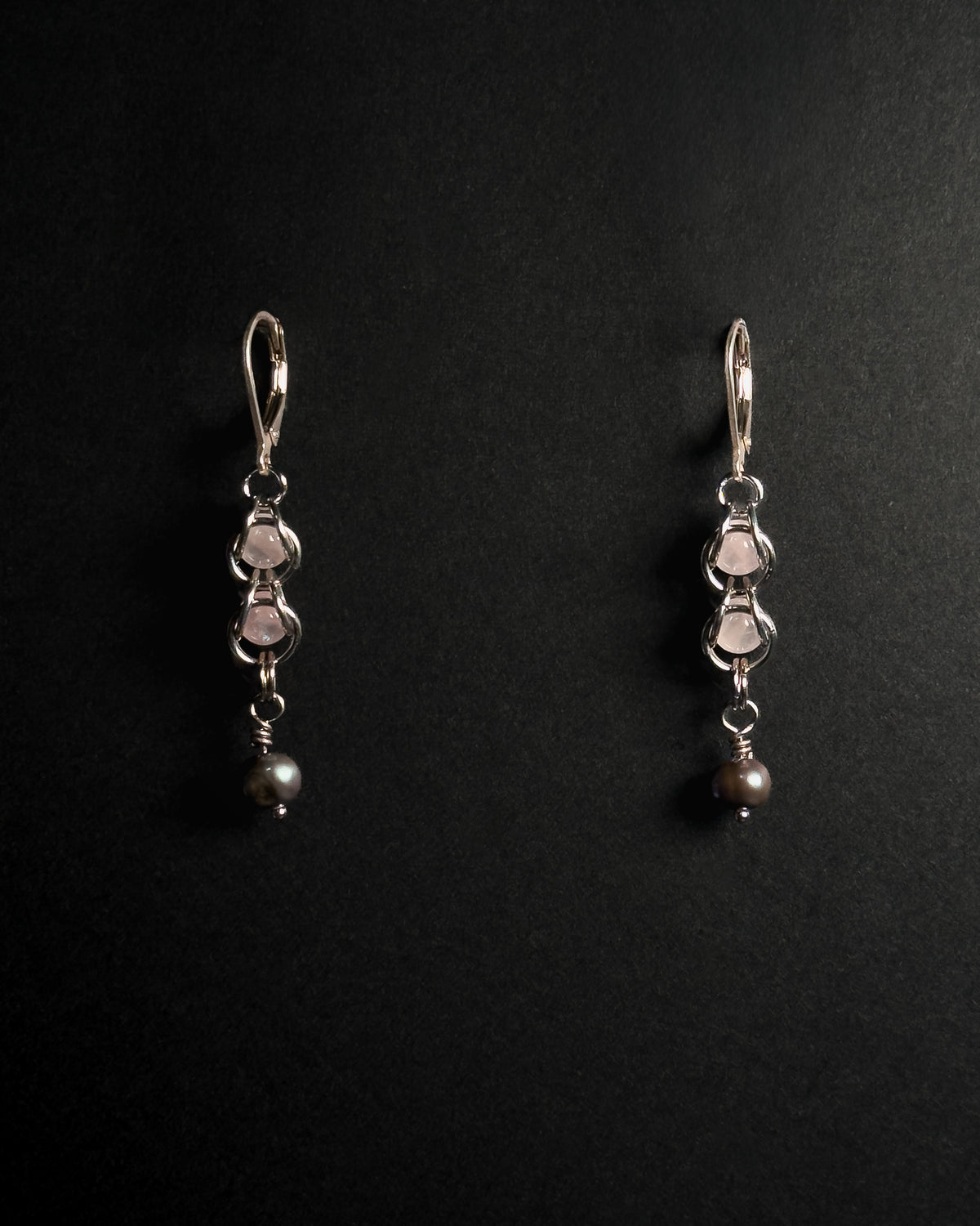 A pair of silver dangle earrings with rose quartz crystal capture bead and dark gray pearl drops, displayed on a black background .