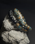 Two 8mm lake blue variscite beaded bracelets displayed on a textured gray rock against a dark background. One bracelet is made of matte black lava stones, and the other is made of full 8mm lake blue variscite with adjustable sliding knot closure.