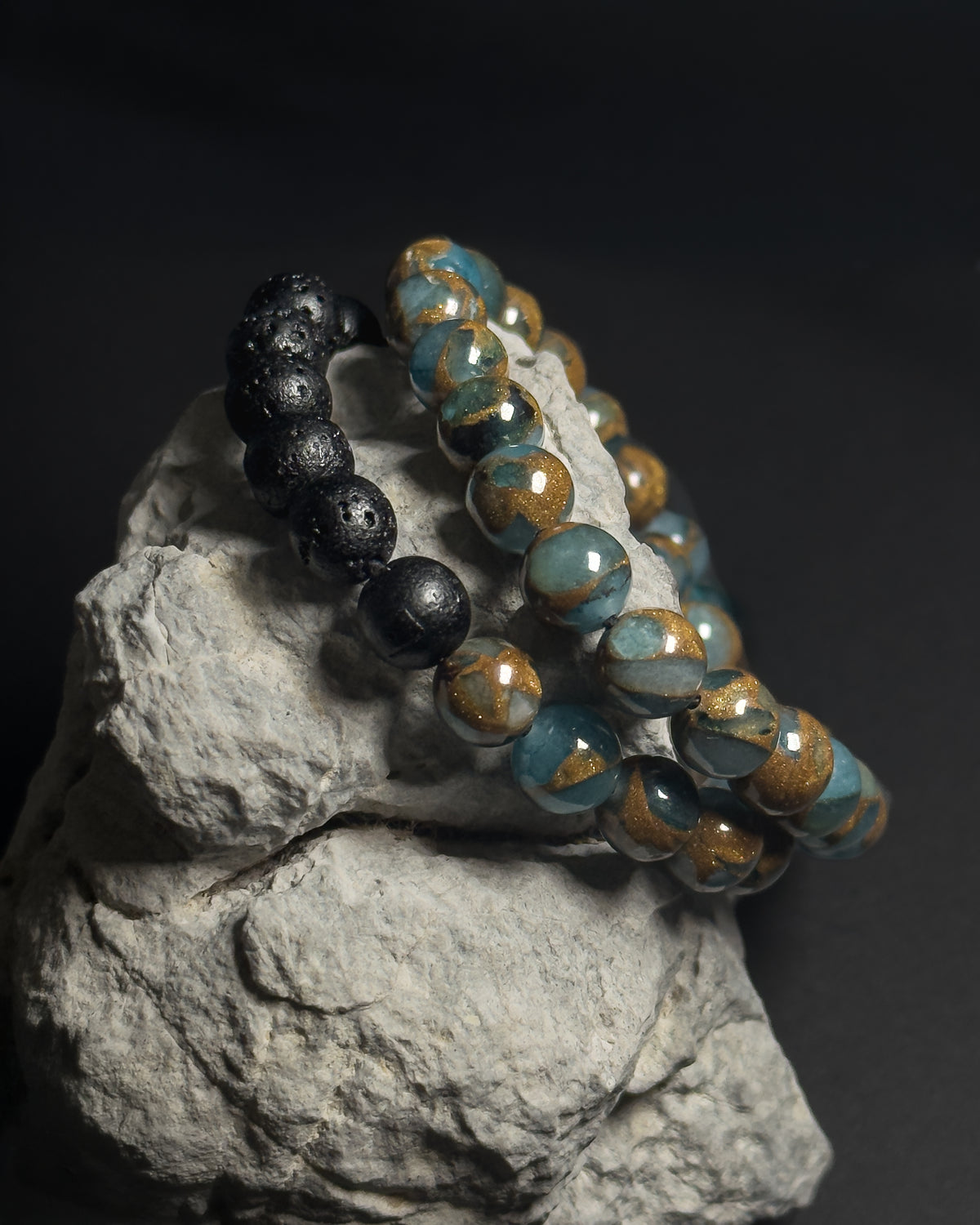 Two 8mm lake blue variscite beaded bracelets displayed on a textured gray rock against a dark background. One bracelet is made of matte black lava stones, and the other is made of full 8mm lake blue variscite with adjustable sliding knot closure.