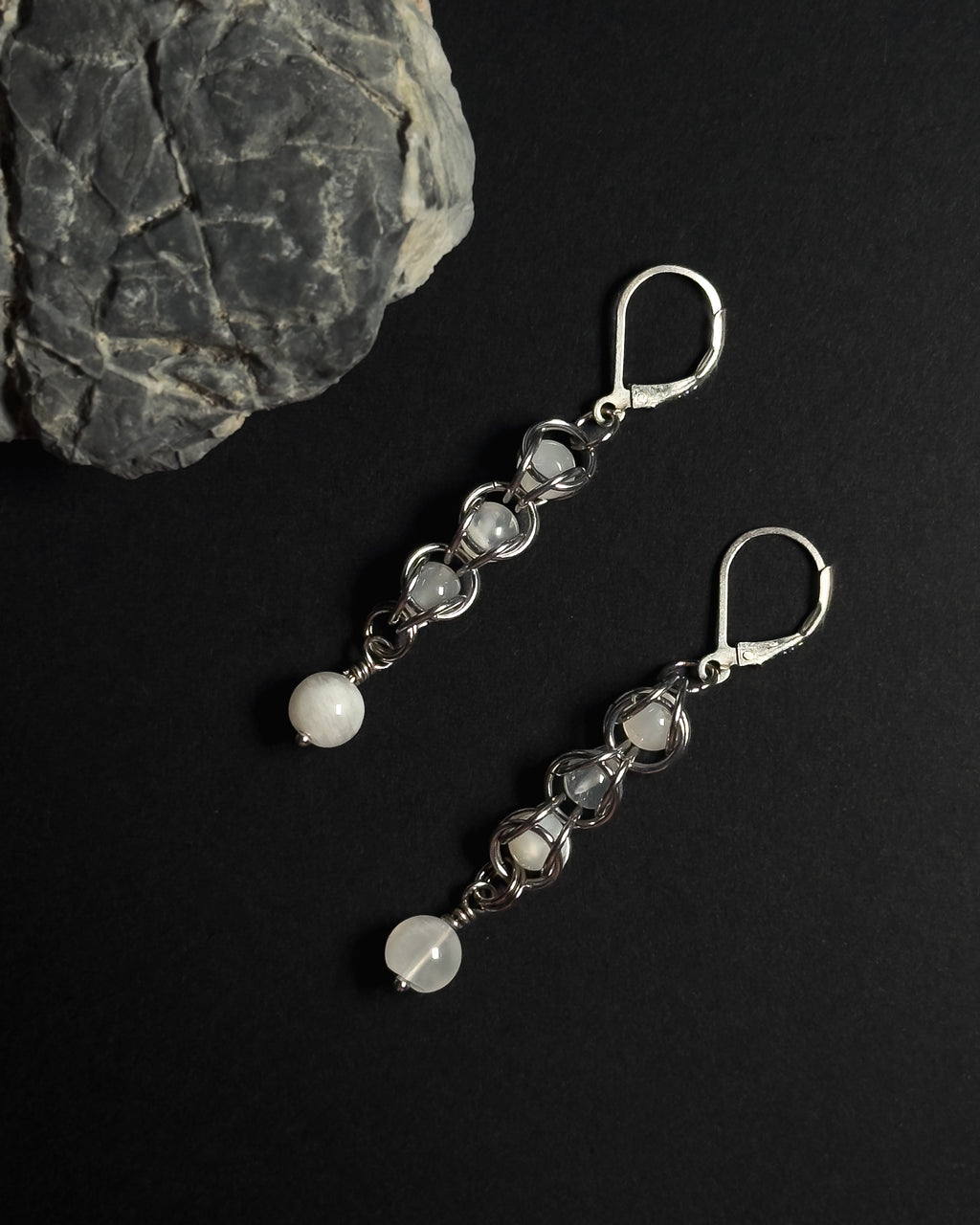 A pair of silver drop earrings with white round beads, displayed on a black background next to a gray rock.