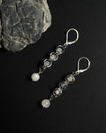 A pair of silver drop earrings with white round beads, displayed on a black background next to a gray rock.
