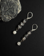A pair of silver drop earrings with white round beads, displayed on a black background next to a gray rock.