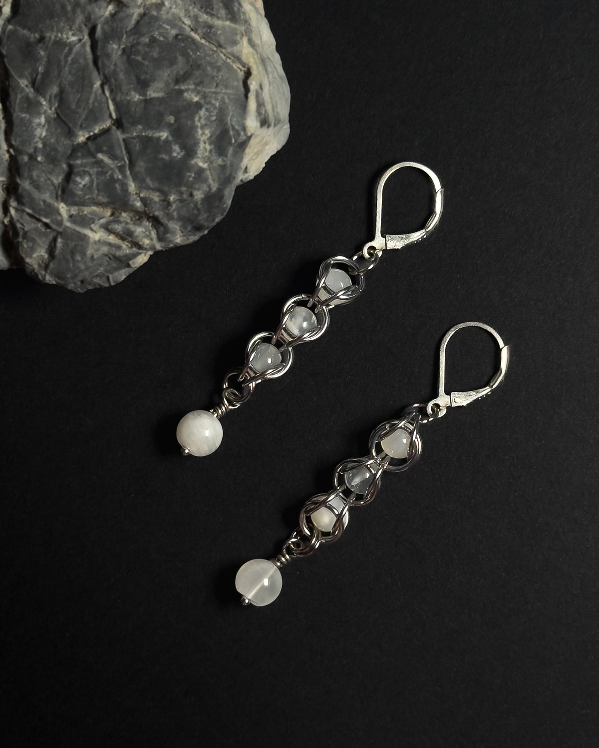 A pair of silver drop earrings with white round beads, displayed on a black background next to a gray rock.