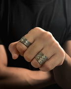 A close-up of a person's hand wearing two wide Solid stainless steel spoon ring with engraved geometric patterns, set against a dark black shirt background.