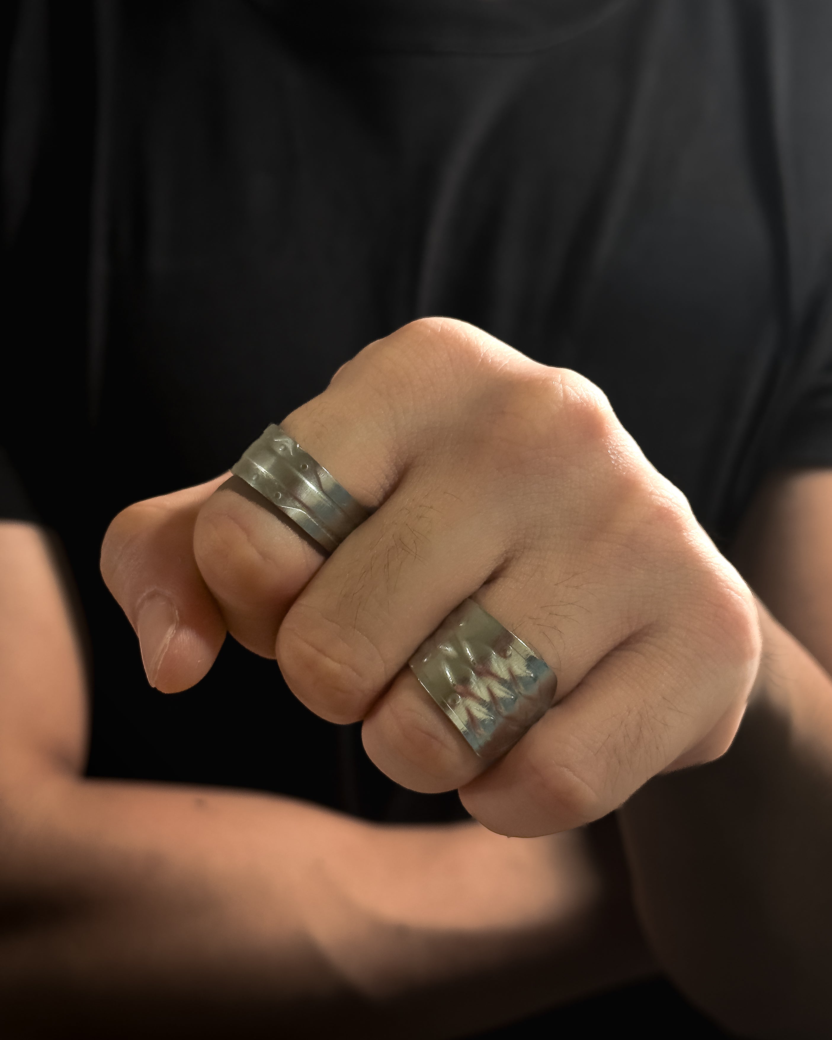 A close-up of a person's hand wearing two wide Solid stainless steel spoon ring with engraved geometric patterns, set against a dark black shirt background.