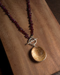 A beaded necklace with reddish purple garnet-like chips and a large oval gold rutilated quartz pendant, displayed on a wooden surface.