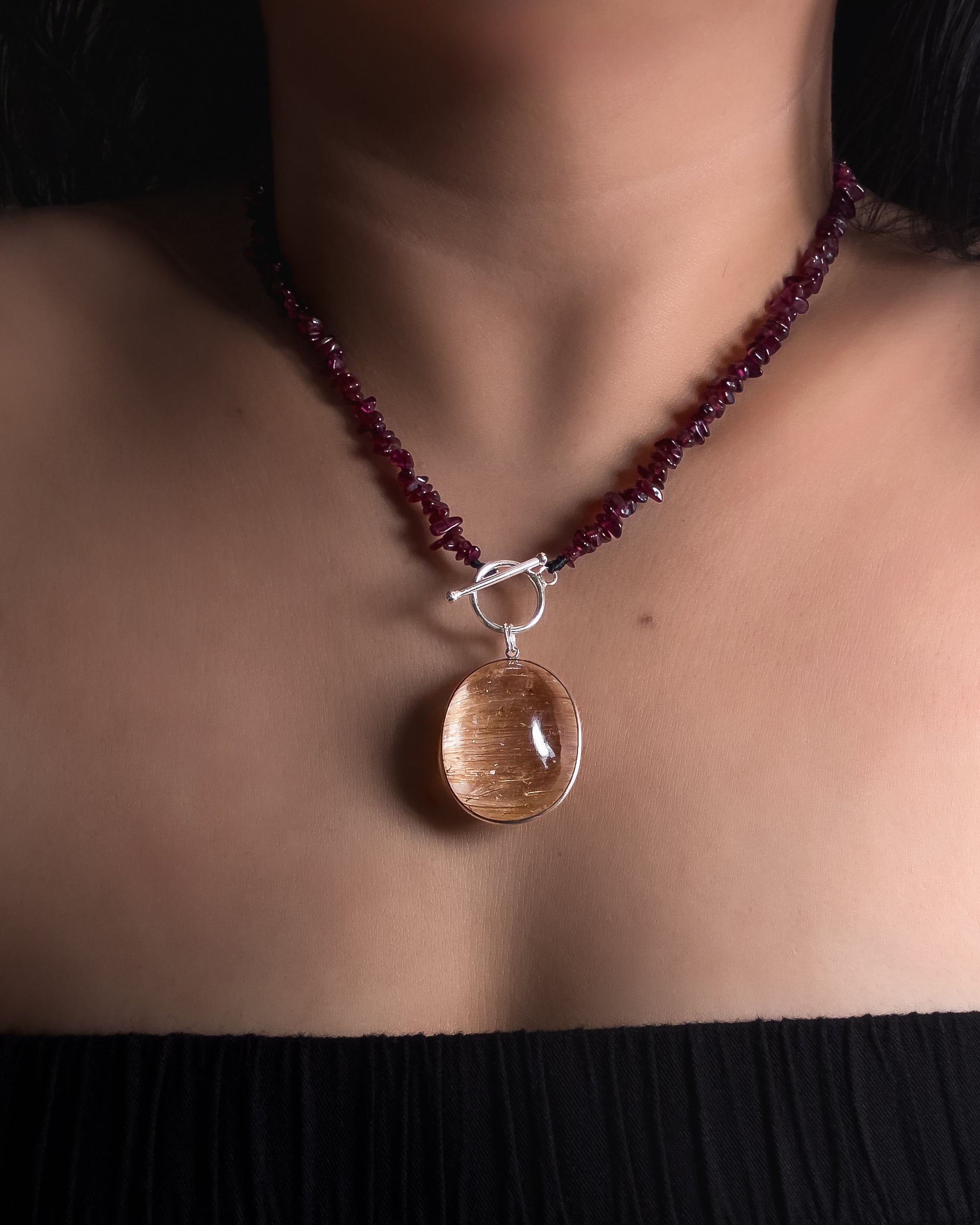 A close-up of a person's neck and chest wearing a necklace with a dark reddish purple garnet beaded necklace, a silver toggle clasp, and a large oval golden rutilated quartz natural gemstone pendant.