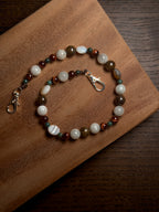 A beaded phone charm with white, brown, green, and reddish-brown beads and silver-tone stainless steel lobster clasp, displayed on a wooden board.