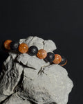 A beaded bracelet with alternating natural sunstone and black lava beads, displayed on a textured gray rock against a solid black background.