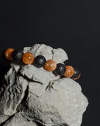 A beaded bracelet with alternating natural sunstone and black lava beads, displayed on a textured gray rock against a solid black background.