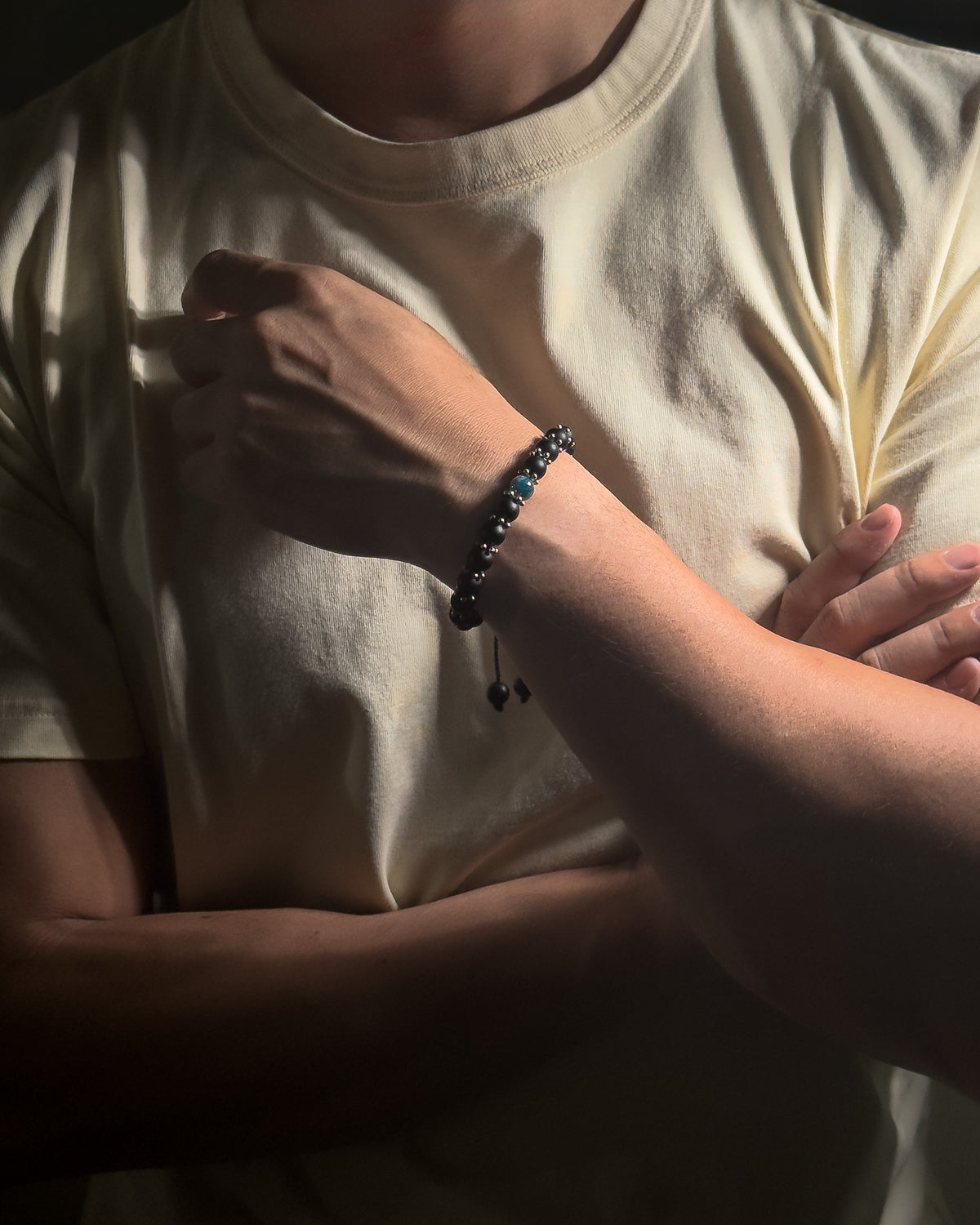 A close-up lifestyle shot of a person wearing a dark matte onyx beaded bracelet with stainless steel spacer beads, featuring blue apatite, wearing a cream-colored t-shirt under dramatic, low-key lighting