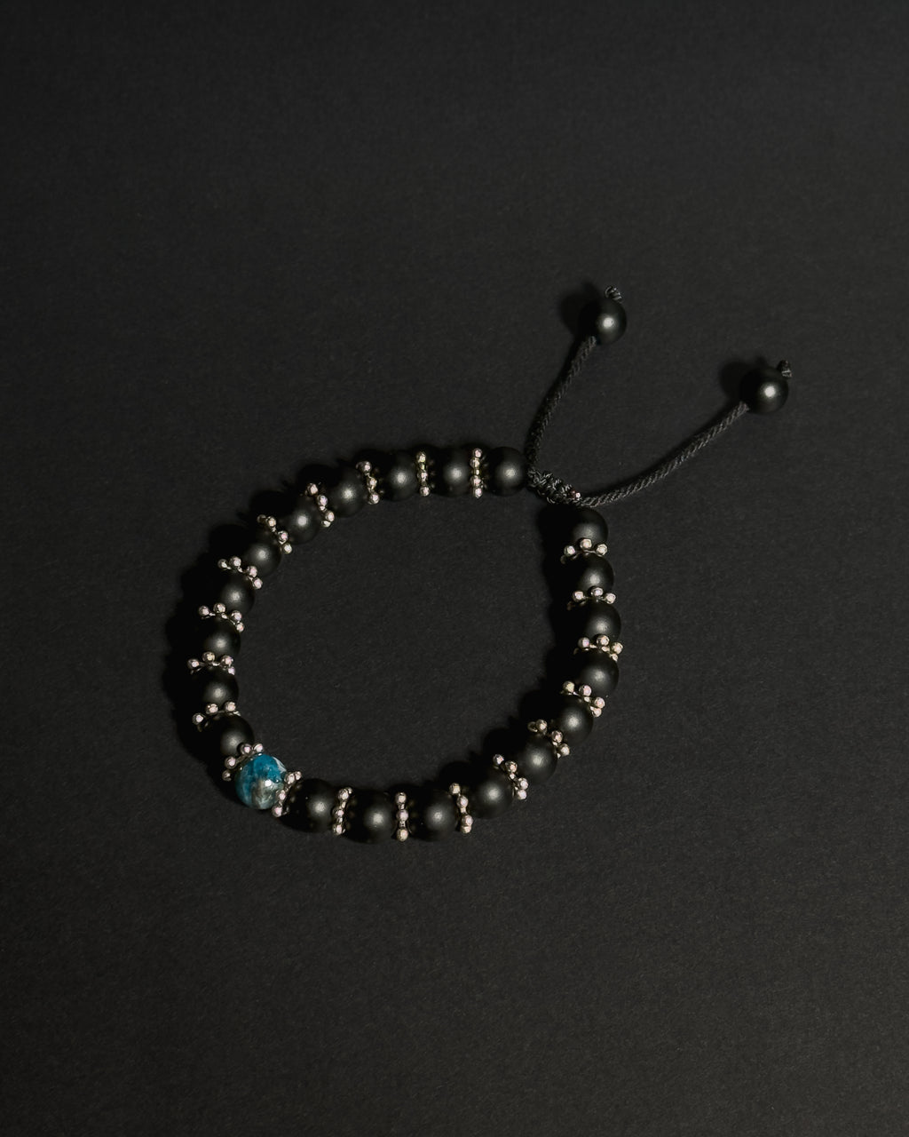 A beaded adjustable bracelet with matte black onyx beads, small metallic spacer beads, and a single blue apatite bead, displayed on a dark background.