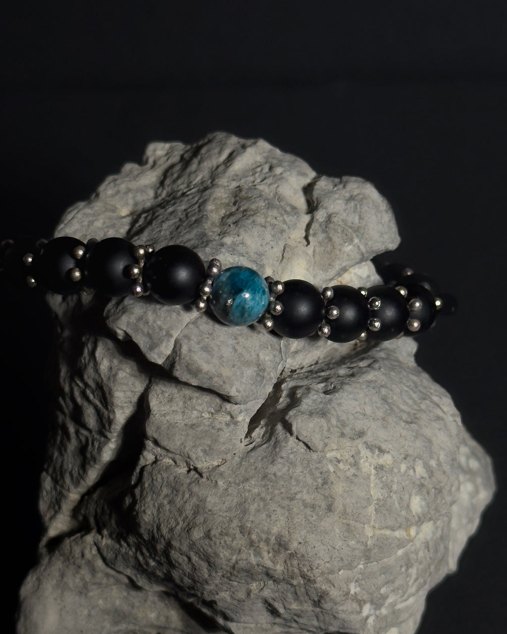 A beaded bracelet with matte black beads, small silver spacer beads, and one prominent round apatite-blue bead, displayed on a textured gray rock against a dark background.