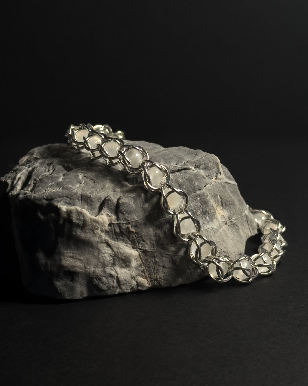 A silver-toned capture bead bracelet with natural white moonstone gemstones draped over a textured gray rock against a solid black background.