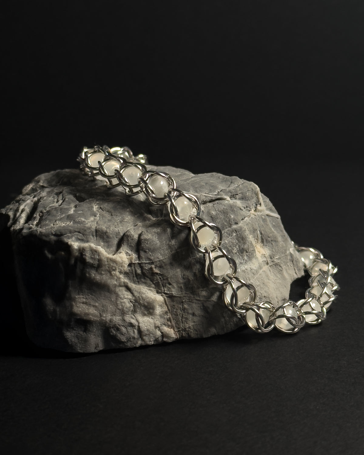 A silver-toned capture bead bracelet with natural white moonstone gemstones draped over a textured gray rock against a solid black background.