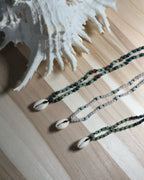 Three natural stone beaded necklaces with shell pendants on a wooden surface next to a large shell.