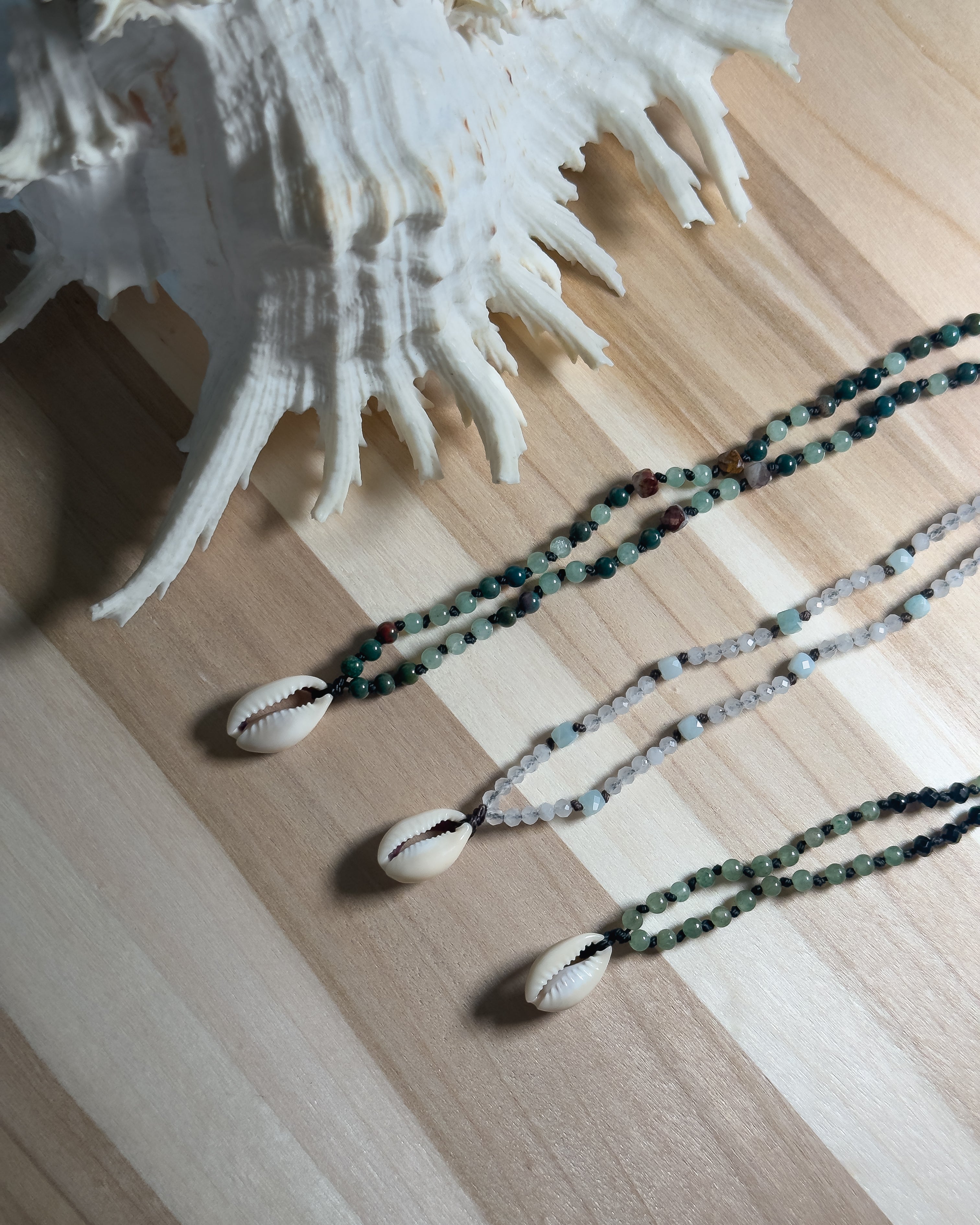 Three natural stone beaded necklaces with shell pendants on a wooden surface next to a large shell.