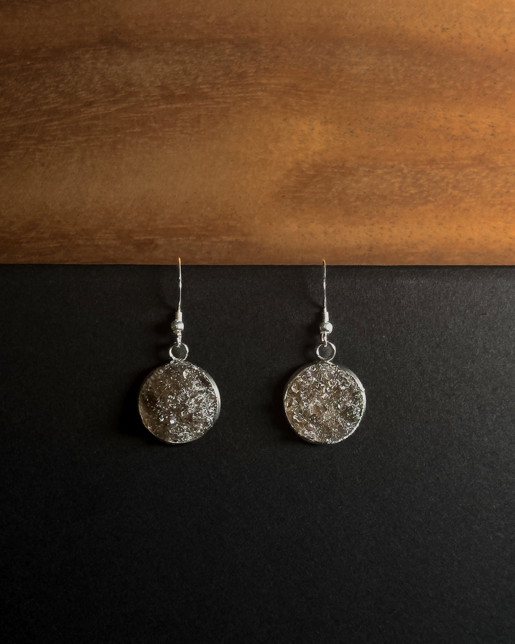 A pair of silver-toned dangle earrings with textured, crushed smokey quartz inlay in round disc pendants, displayed on a split black and wood background.
