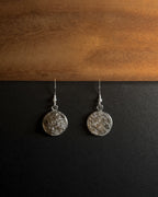 A pair of silver-toned dangle earrings with textured, crushed smokey quartz inlay in round disc pendants, displayed on a split black and wood background.