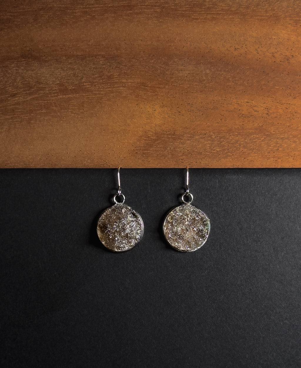 A pair of silver-toned dangle earrings with textured, crushed smokey quartz inlay in round disc pendants, displayed on a split black and wood background.