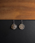 A pair of silver-toned dangle earrings with textured, crushed smokey quartz inlay in round disc pendants, displayed on a split black and wood background.