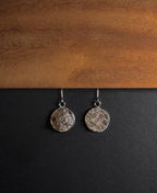 A pair of silver-toned dangle earrings with textured, crushed smokey quartz inlay in round disc pendants, displayed on a split black and wood background.