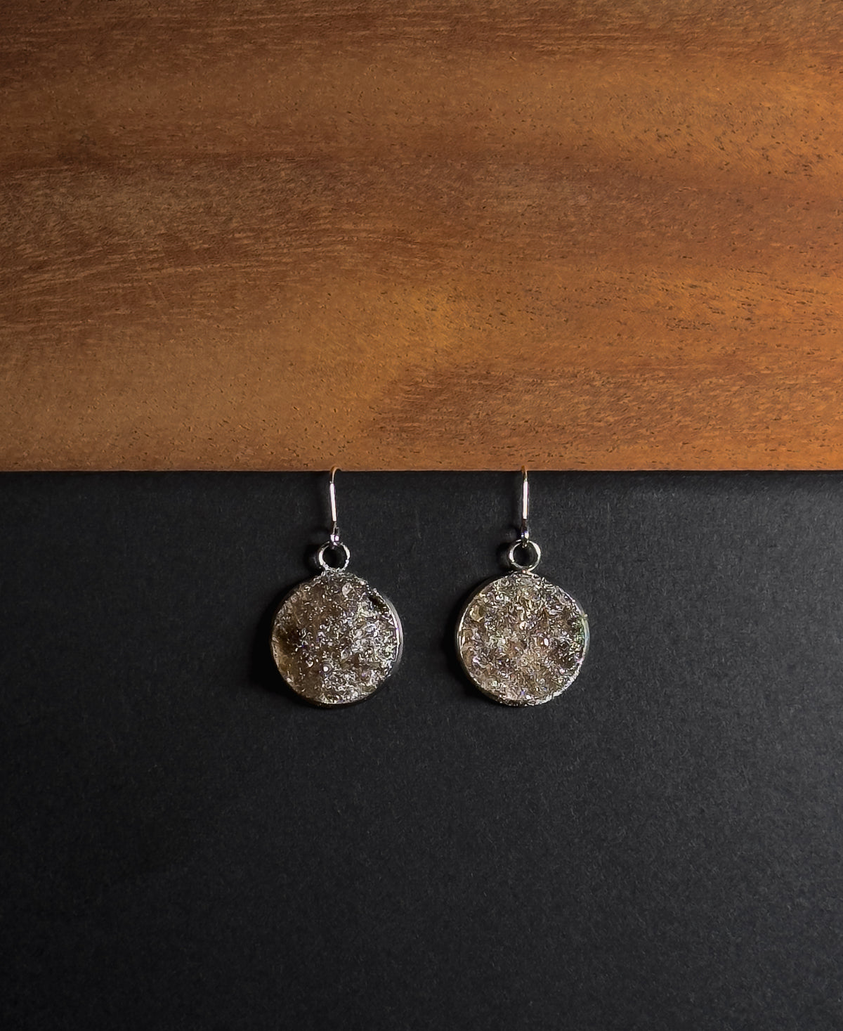 A pair of silver-toned dangle earrings with textured, crushed smokey quartz inlay in round disc pendants, displayed on a split black and wood background.
