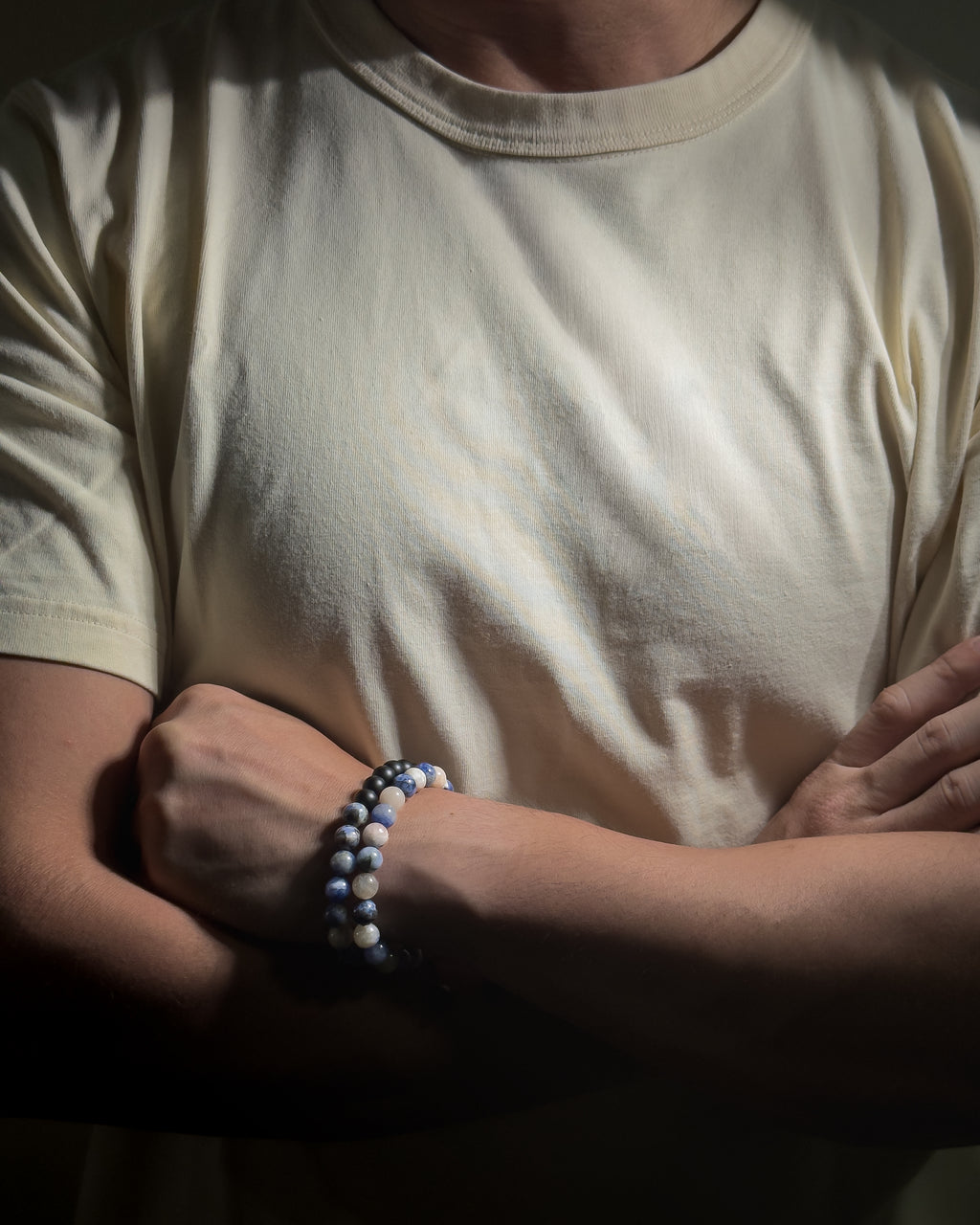 A close-up of a person wearing a cream-colored t-shirt and beaded bracelet on their wrist, with their arms crossed in a dimly lit setting.
