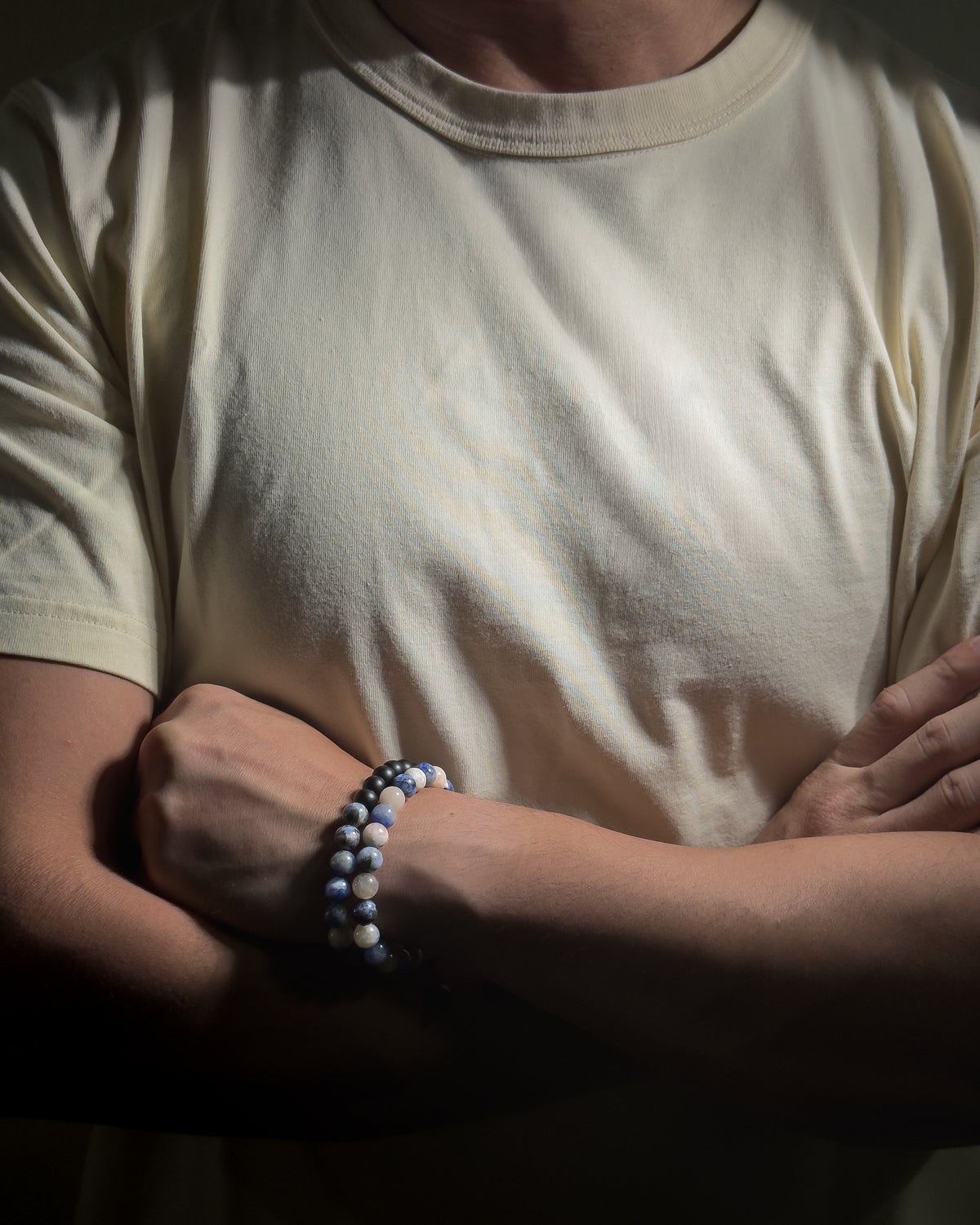 A close-up of a person wearing a cream-colored t-shirt and beaded bracelet on their wrist, with their arms crossed in a dimly lit setting.