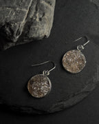 A pair of round silver-toned dangle earrings with rough, textured crushed smokey quartz stone inlays, displayed on a dark slate surface next to a larger rock.