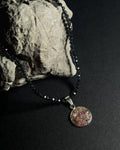 A beaded necklace with dark faceted spinel beads and a round pendant containing multicolour natural crushed stones, rainbow tourmaline, displayed on a dark surface next to a textured stone.