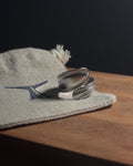 Solid stainless steel spiral spoon ring resting on a beige woven fabric pouch with a dark blue logo, placed on a wooden surface against a dark background.