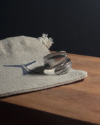 Solid stainless steel spiral spoon ring resting on a beige woven fabric pouch with a dark blue logo, placed on a wooden surface against a dark background.