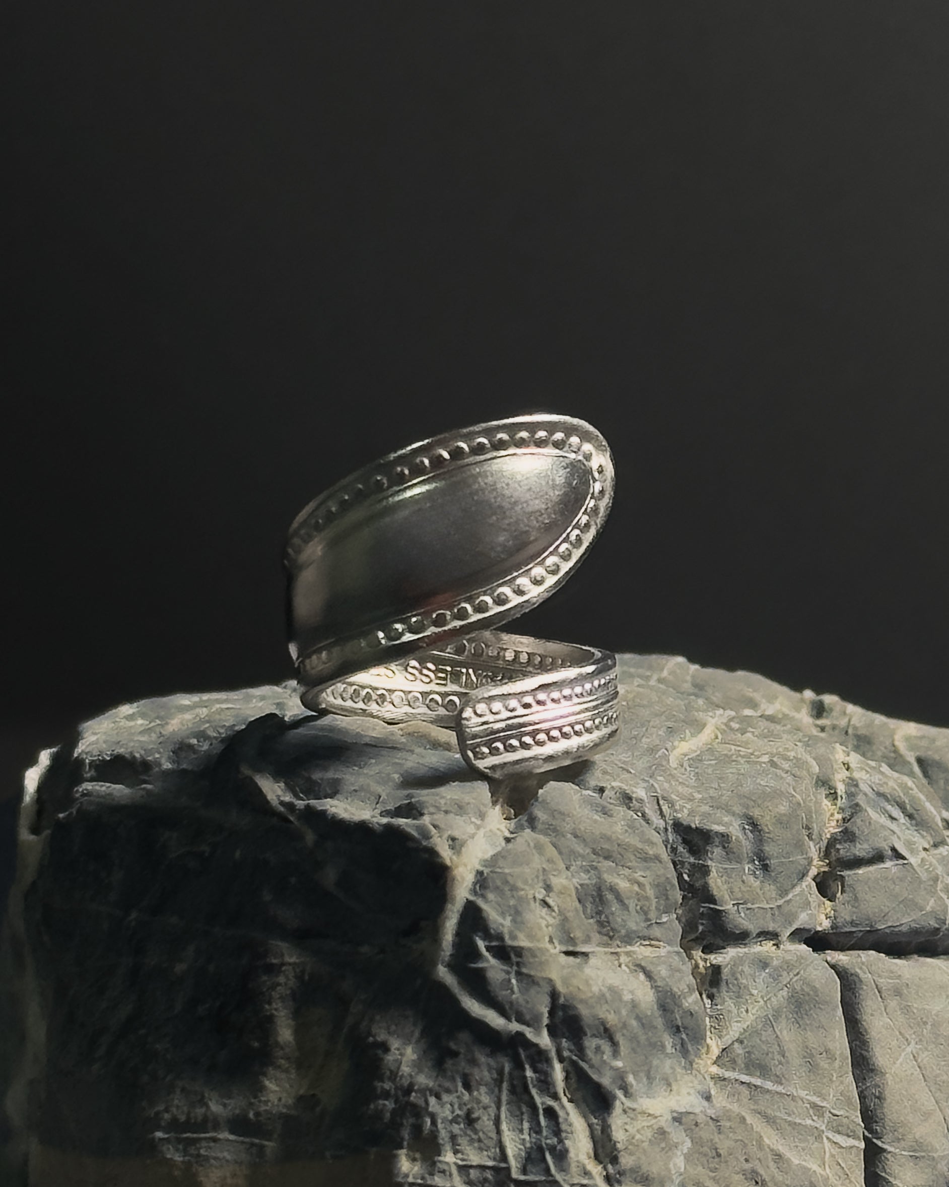 Solid stainless steel spoon ring with a textured, stud-like in a wrap finish, displayed on a textured dark gray rock against a black background.