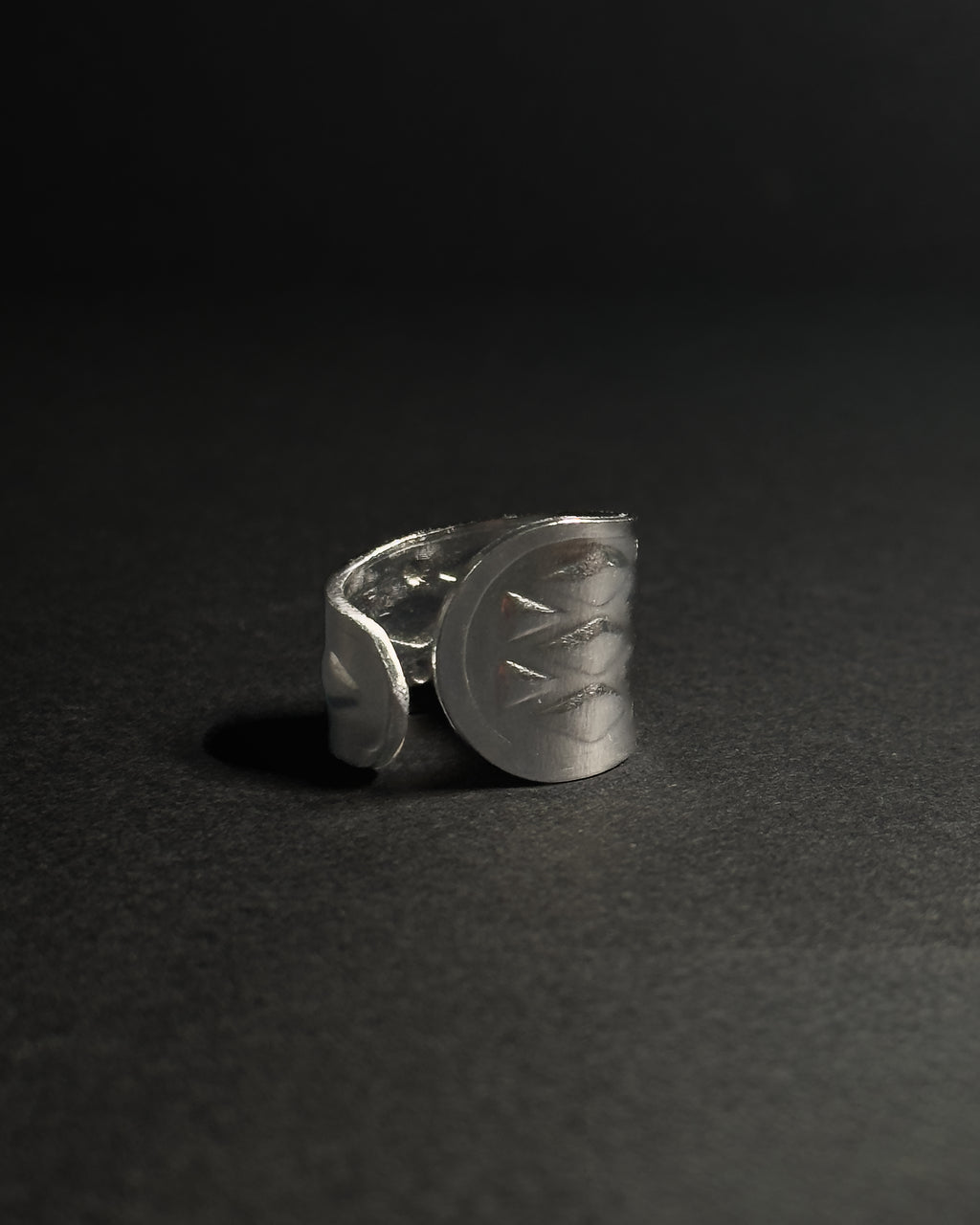 Solid stainless steel spoon ring with a wide, shield-like face featuring engraved geometric patterns, photographed on a dark textured surface against a dark background.