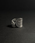 Solid stainless steel spoon ring with a wide, shield-like face featuring engraved geometric patterns, photographed on a dark textured surface against a dark background.