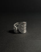 Solid stainless steel spoon ring with a wide, shield-like face featuring engraved geometric patterns, photographed on a dark textured surface against a dark background.