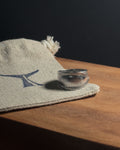 A polishedSolid stainless steel spoon ring with decorative stud-like detailing on the band, resting on a beige woven linen fabric pouch with a blue logo, placed on a wooden surface against a dark background.