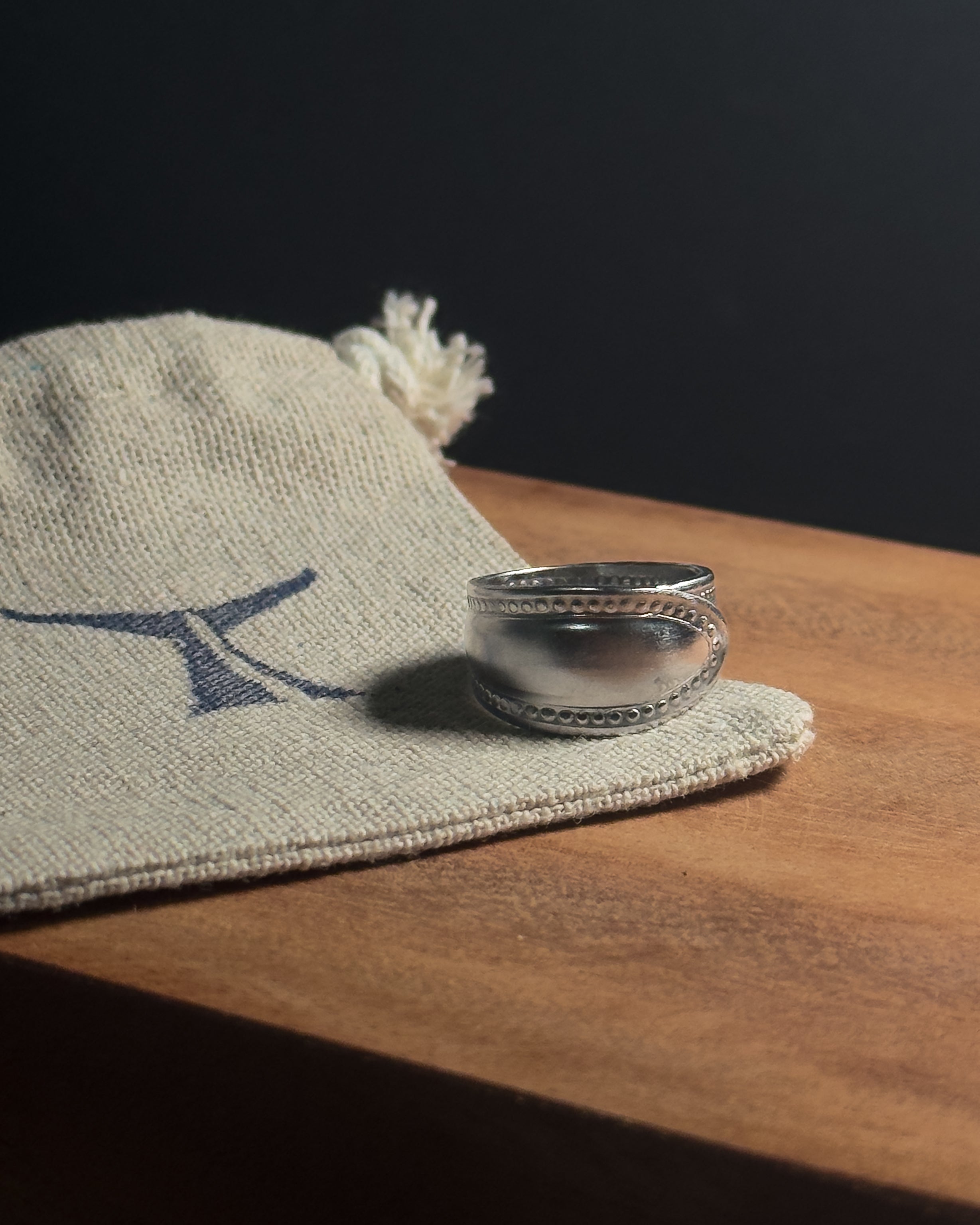 A polishedSolid stainless steel spoon ring with decorative stud-like detailing on the band, resting on a beige woven linen fabric pouch with a blue logo, placed on a wooden surface against a dark background.